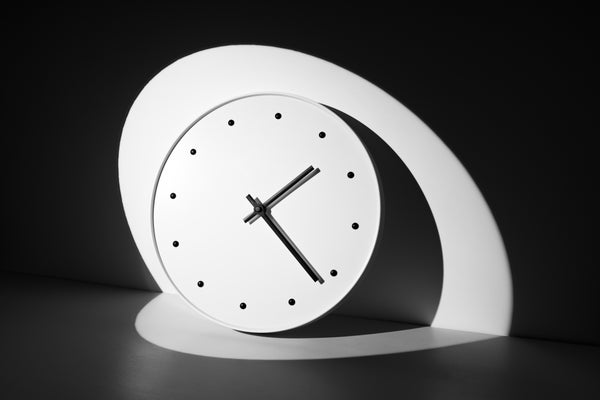 A high angled spotlight focused on a clock leaning on a wall in a dark space, casting a sharp shadow onto the wall and surface behind the clock